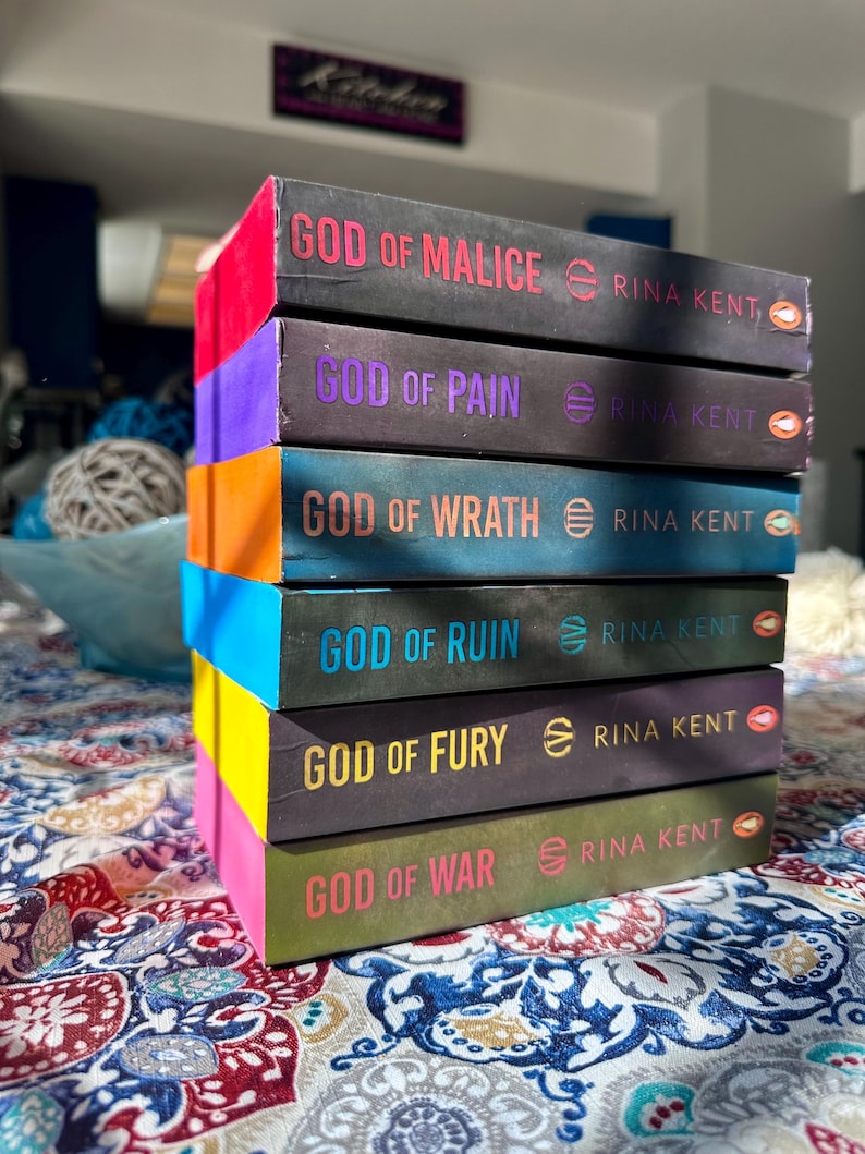 A complete 6-book set of Rina Kent's Legacy of Gods series featuring custom sprayed edges. The book edges are airbrushed in a solid rainbow gradient of red, purple, orange, blue, yellow, and pink to perfectly match the original covers. The books are stacked on a white shelf in a minimalist "BookTok" aesthetic style.