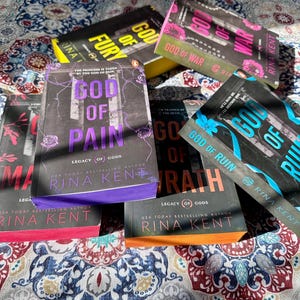 A complete 6-book set of Rina Kent's Legacy of Gods series featuring custom sprayed edges. The book edges are airbrushed in a solid rainbow gradient of red, purple, orange, blue, yellow, and pink to perfectly match the original covers. The books are stacked on a white shelf in a minimalist "BookTok" aesthetic style.