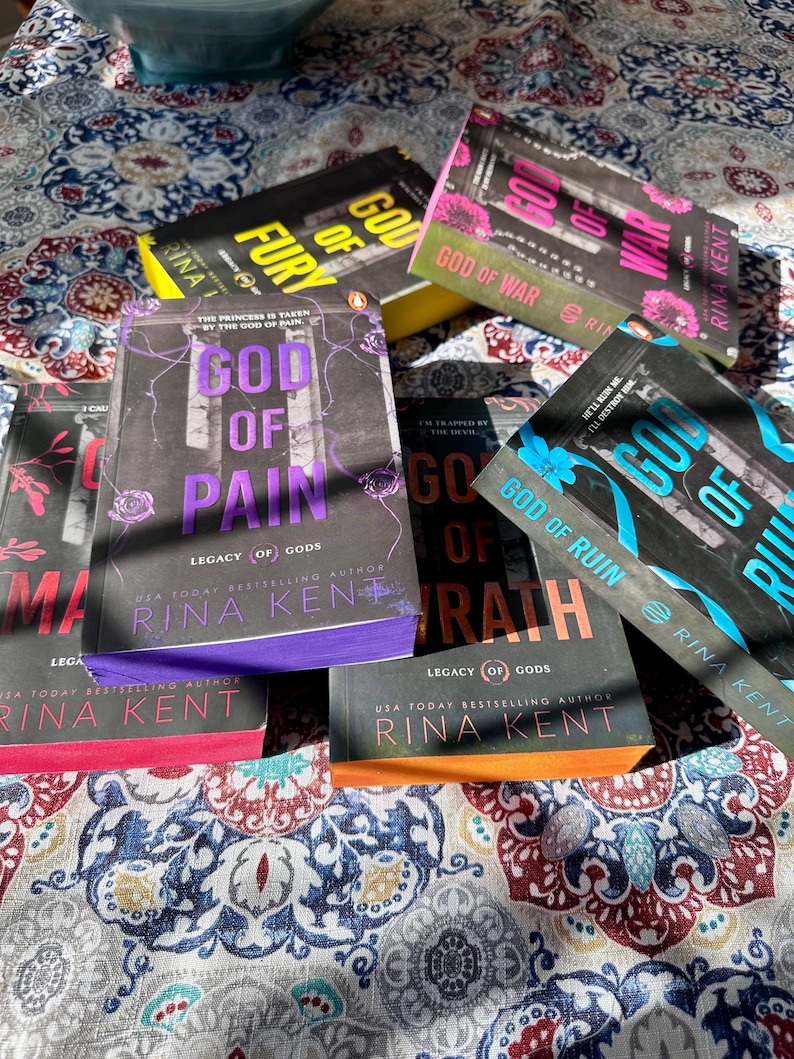 A complete 6-book set of Rina Kent's Legacy of Gods series featuring custom sprayed edges. The book edges are airbrushed in a solid rainbow gradient of red, purple, orange, blue, yellow, and pink to perfectly match the original covers. The books are stacked on a white shelf in a minimalist "BookTok" aesthetic style.