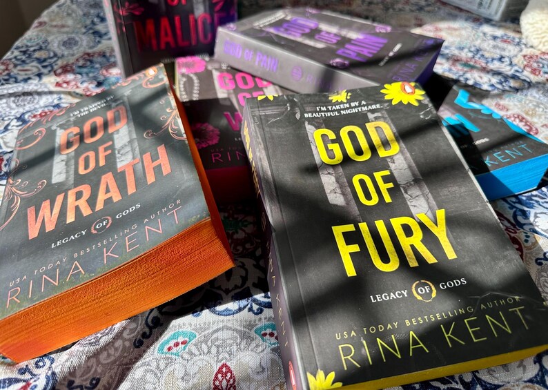 A complete 6-book set of Rina Kent's Legacy of Gods series featuring custom sprayed edges. The book edges are airbrushed in a solid rainbow gradient of red, purple, orange, blue, yellow, and pink to perfectly match the original covers. The books are stacked on a white shelf in a minimalist "BookTok" aesthetic style.