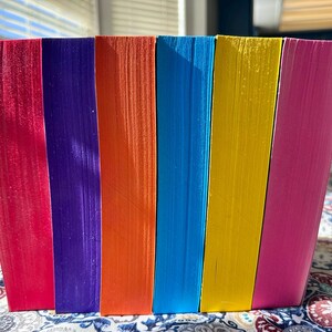 A complete 6-book set of Rina Kent's Legacy of Gods series featuring custom sprayed edges. The book edges are airbrushed in a solid rainbow gradient of red, purple, orange, blue, yellow, and pink to perfectly match the original covers. The books are stacked on a white shelf in a minimalist "BookTok" aesthetic style.