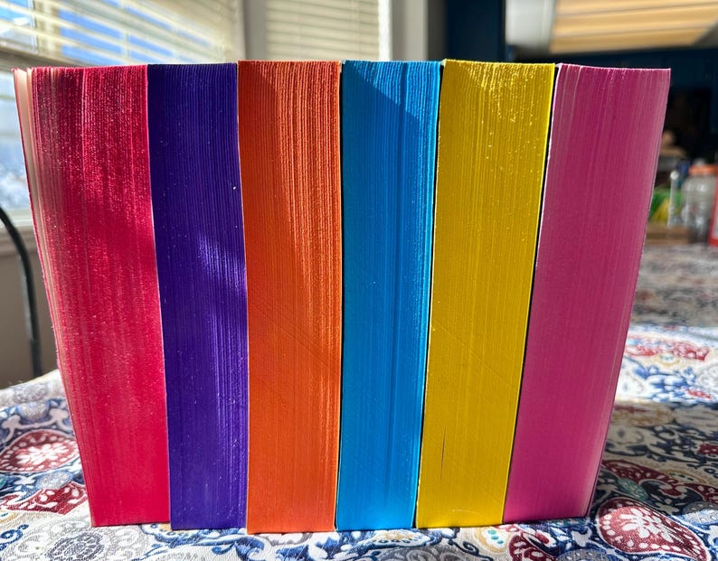 A complete 6-book set of Rina Kent's Legacy of Gods series featuring custom sprayed edges. The book edges are airbrushed in a solid rainbow gradient of red, purple, orange, blue, yellow, and pink to perfectly match the original covers. The books are stacked on a white shelf in a minimalist "BookTok" aesthetic style.