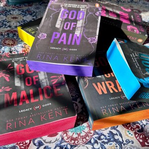A complete 6-book set of Rina Kent's Legacy of Gods series featuring custom sprayed edges. The book edges are airbrushed in a solid rainbow gradient of red, purple, orange, blue, yellow, and pink to perfectly match the original covers. The books are stacked on a white shelf in a minimalist "BookTok" aesthetic style.