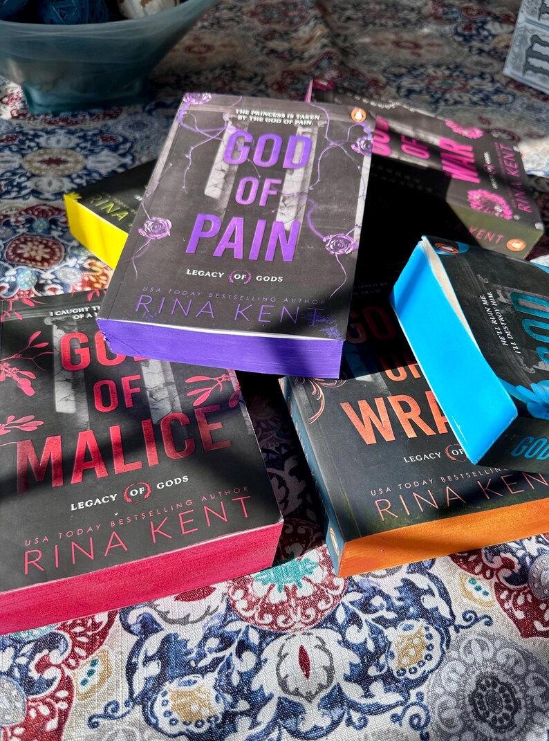 A complete 6-book set of Rina Kent's Legacy of Gods series featuring custom sprayed edges. The book edges are airbrushed in a solid rainbow gradient of red, purple, orange, blue, yellow, and pink to perfectly match the original covers. The books are stacked on a white shelf in a minimalist "BookTok" aesthetic style.