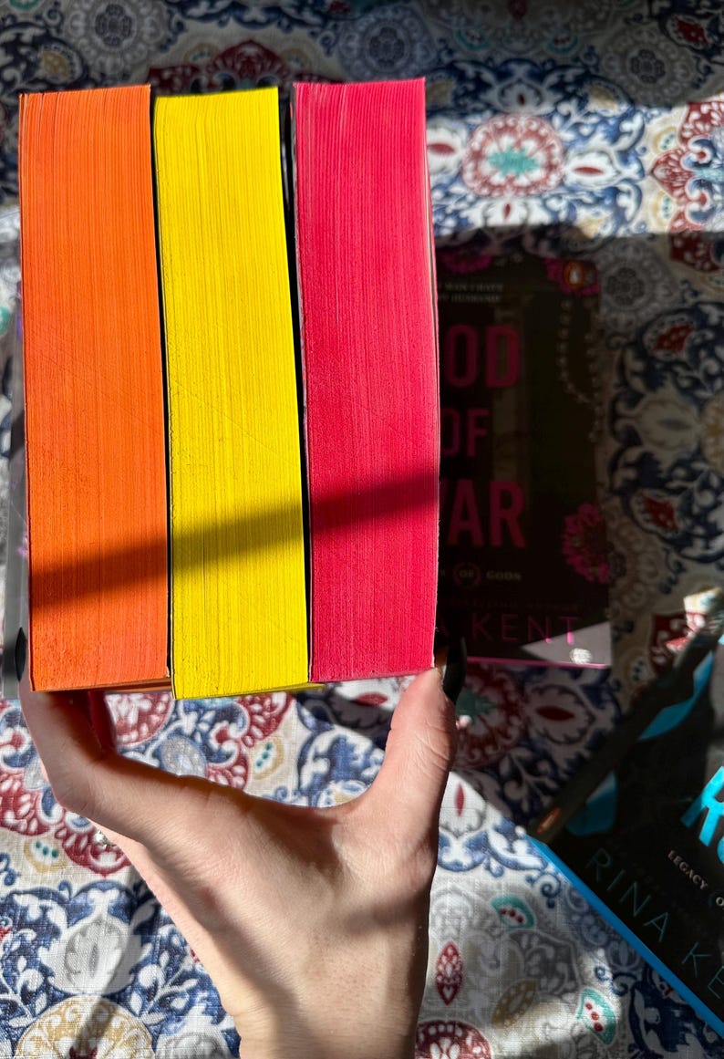 A complete 6-book set of Rina Kent's Legacy of Gods series featuring custom sprayed edges. The book edges are airbrushed in a solid rainbow gradient of red, purple, orange, blue, yellow, and pink to perfectly match the original covers. The books are stacked on a white shelf in a minimalist "BookTok" aesthetic style.