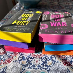 A complete 6-book set of Rina Kent's Legacy of Gods series featuring custom sprayed edges. The book edges are airbrushed in a solid rainbow gradient of red, purple, orange, blue, yellow, and pink to perfectly match the original covers. The books are stacked on a white shelf in a minimalist "BookTok" aesthetic style.