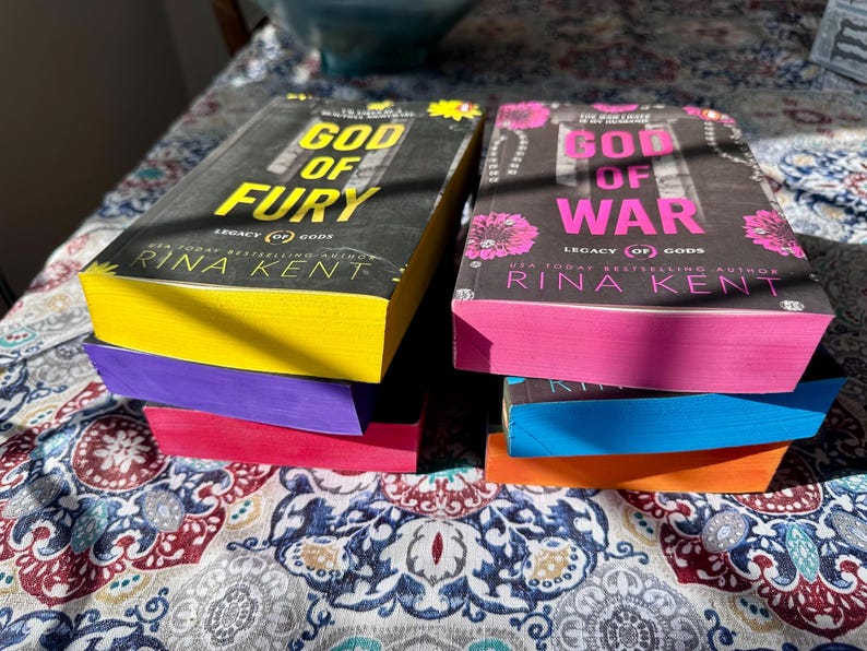 A complete 6-book set of Rina Kent's Legacy of Gods series featuring custom sprayed edges. The book edges are airbrushed in a solid rainbow gradient of red, purple, orange, blue, yellow, and pink to perfectly match the original covers. The books are stacked on a white shelf in a minimalist "BookTok" aesthetic style.