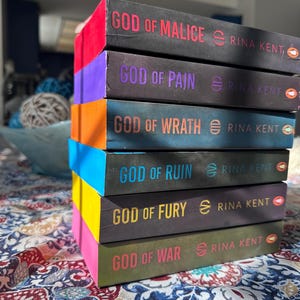 A complete 6-book set of Rina Kent's Legacy of Gods series featuring custom sprayed edges. The book edges are airbrushed in a solid rainbow gradient of red, purple, orange, blue, yellow, and pink to perfectly match the original covers. The books are stacked on a white shelf in a minimalist "BookTok" aesthetic style.