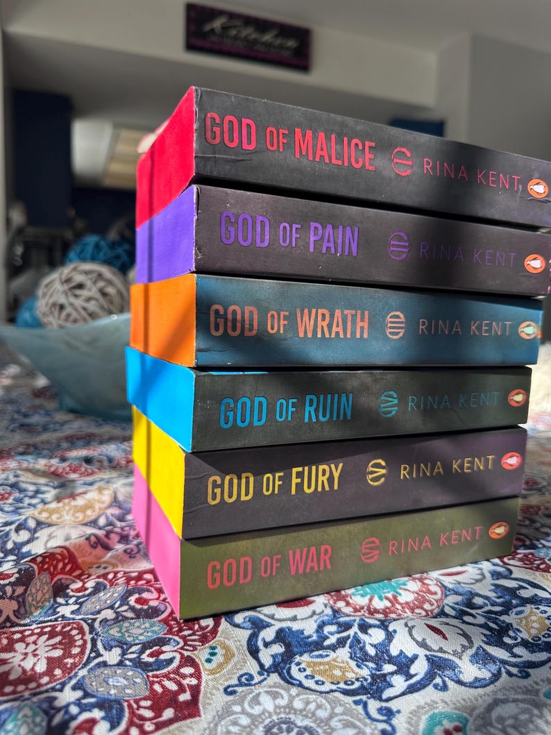 A complete 6-book set of Rina Kent's Legacy of Gods series featuring custom sprayed edges. The book edges are airbrushed in a solid rainbow gradient of red, purple, orange, blue, yellow, and pink to perfectly match the original covers. The books are stacked on a white shelf in a minimalist "BookTok" aesthetic style.