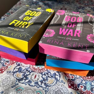 A complete 6-book set of Rina Kent's Legacy of Gods series featuring custom sprayed edges. The book edges are airbrushed in a solid rainbow gradient of red, purple, orange, blue, yellow, and pink to perfectly match the original covers. The books are stacked on a white shelf in a minimalist "BookTok" aesthetic style.