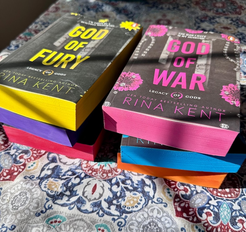 A complete 6-book set of Rina Kent's Legacy of Gods series featuring custom sprayed edges. The book edges are airbrushed in a solid rainbow gradient of red, purple, orange, blue, yellow, and pink to perfectly match the original covers. The books are stacked on a white shelf in a minimalist "BookTok" aesthetic style.