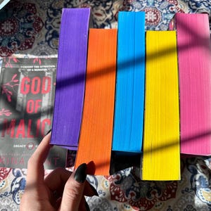 May include: A collection of colorful books with bright edges. The spines are purple, orange, blue, yellow, and pink. The cover of one book reads "God of Malice" with the text "Legacy of Gods" and the author's name.