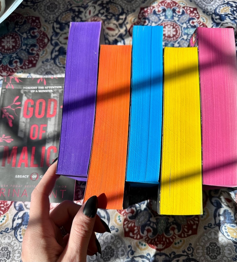 May include: A collection of colorful books with bright edges. The spines are purple, orange, blue, yellow, and pink. The cover of one book reads "God of Malice" with the text "Legacy of Gods" and the author's name.