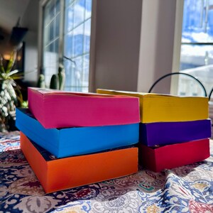 A complete 6-book set of Rina Kent's Legacy of Gods series featuring custom sprayed edges. The book edges are airbrushed in a solid rainbow gradient of red, purple, orange, blue, yellow, and pink to perfectly match the original covers. The books are stacked on a white shelf in a minimalist "BookTok" aesthetic style.