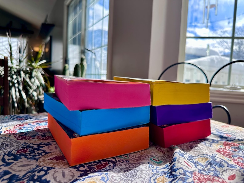 A complete 6-book set of Rina Kent's Legacy of Gods series featuring custom sprayed edges. The book edges are airbrushed in a solid rainbow gradient of red, purple, orange, blue, yellow, and pink to perfectly match the original covers. The books are stacked on a white shelf in a minimalist "BookTok" aesthetic style.