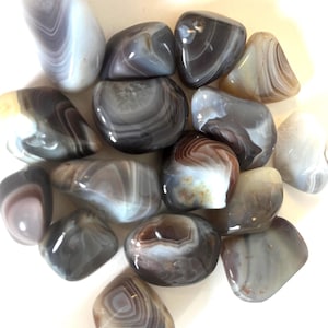 May include: A selection of polished agate stones in various shapes and sizes. The stones display a range of colours, including grey, white, brown, and tan, with banded patterns and swirls. The stones are smooth and glossy.