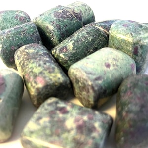 May include: A pile of tumbled stones, mainly green with maroon and purple flecks. The stones are smooth, rounded, and of varying sizes. The overall look is natural and earthy, possibly indicating gemstones or crystals.