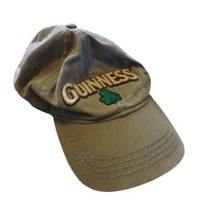 May include: Olive green baseball cap with the word "GUINNESS" embroidered in gold, along with a green shamrock. The cap has a curved brim and a fabric strap closure.