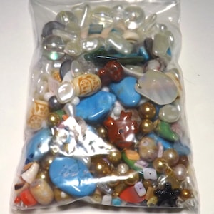May include: A clear plastic bag filled with a variety of beads in different shapes, sizes, and colors. The beads include turquoise, gold, white, red, and iridescent pieces. Some beads have patterns, and others are smooth. The bag is on a white surface.