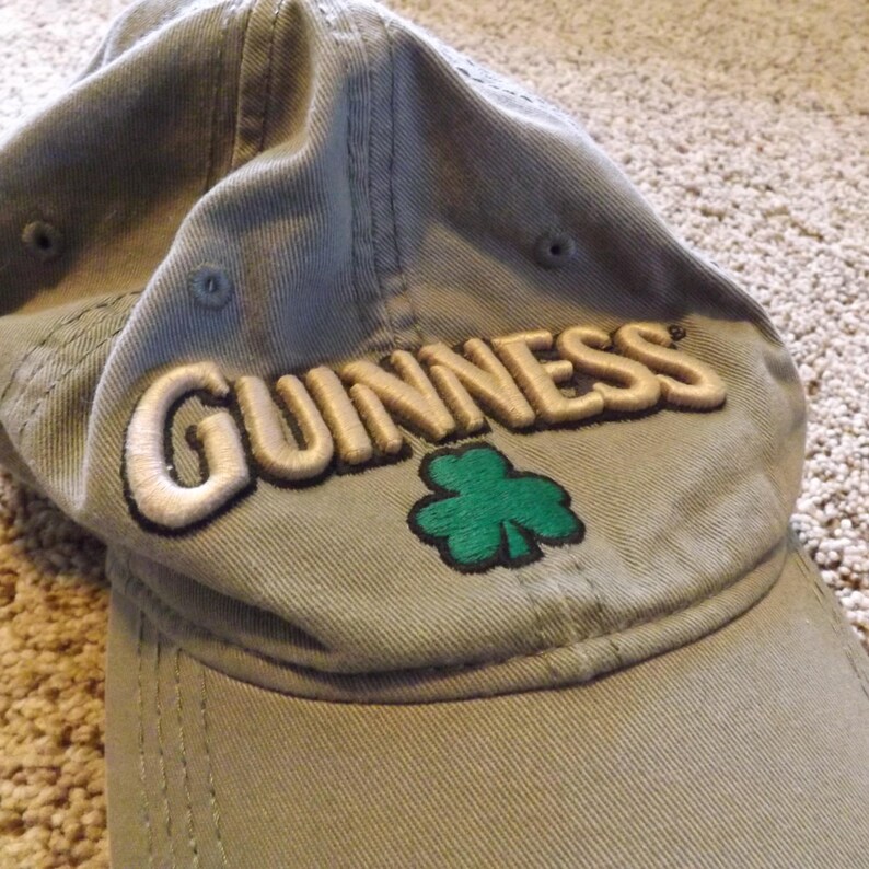 May include: A sage green baseball cap with the word "GUINNESS" embroidered in cream-colored thread. A green shamrock is embroidered below the word. The cap is made of a soft, worn fabric.