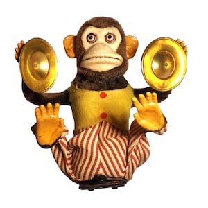 May include: Vintage wind-up toy monkey with a brown head, yellow vest, and red and white striped pants. The monkey holds two golden cymbals and has a painted face with a wide grin. A classic collectible toy.