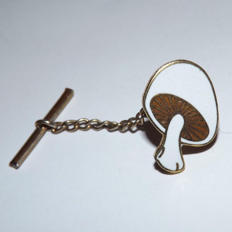 Puede incluir: Un broche de corbata dorado con un dise&ntilde;o de hongo de esmalte blanco. El hongo tiene un sombrero marr&oacute;n con manchas blancas y un tallo blanco.