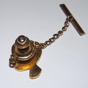 Puede incluir: Un broche de corbata dorado con una cadena y un cierre de barra. El broche de corbata presenta un dise&ntilde;o estilizado de un coraz&oacute;n.
