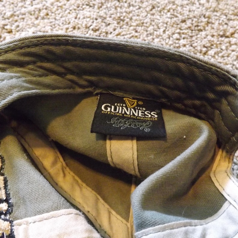 May include: Olive green baseball cap with the Guinness logo and "Official Merchandise" text on a black tag. The cap's interior is tan and white.