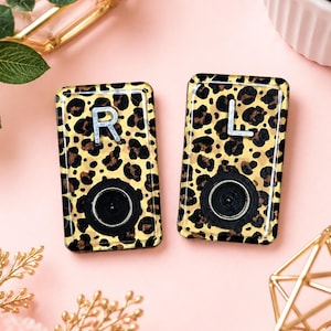 May include: Two rectangular hearing aid covers featuring a glossy leopard print pattern. Each cover displays a silver letter, R on one and L on the other, positioned above a circular black opening. The surface has a smooth, reflective finish.