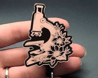 Puede incluir: Un microscopio en blanco y negro con detalles florales en un cordón negro con un portagafete transparente.