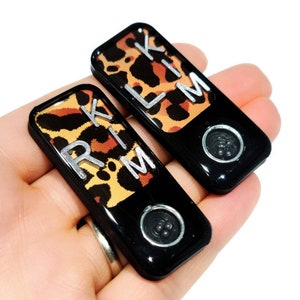 May include: Two black keychains with a leopard print design. The keychains have the letters "RIK" and "LIM" printed on them in silver.