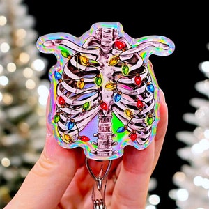 May include: A holographic iridescent charm featuring a skeleton with colorful Christmas lights wrapped around its ribcage. The charm is attached to a silver swivel clip.