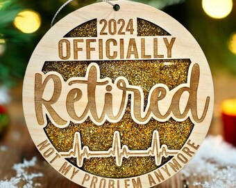 May include: A round wooden Christmas bauble with gold glitter and the text "2024 Officially Retired Not My Problem Anymore".
