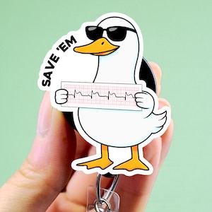 May include: A white duck wearing sunglasses, holding an EKG strip, and the words "SAVE 'EM" on a retractable badge reel. The duck has a yellow beak and feet. The badge reel has a clear plastic strap and a metal clip.