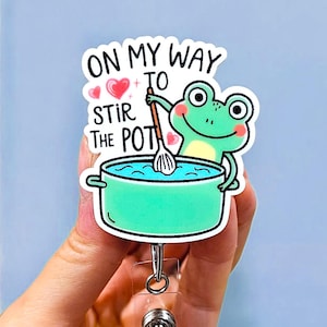 May include: A badge reel featuring a cartoon frog stirring a pot of blue liquid. The text "ON MY WAY TO STIR THE POT" is above the frog. The reel has a clear plastic base and a metal clip.
