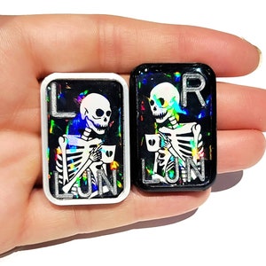 May include: Two resin skeleton keychains with a holographic finish. One keychain is black with white lettering and the other is white with black lettering. Both keychains feature a skeleton holding a coffee cup and the word "LUN" on the bottom.