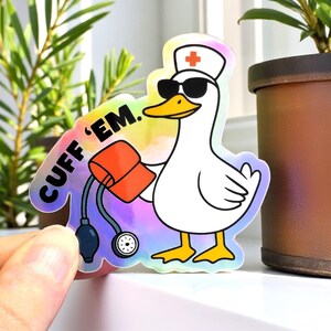 May include: A holographic sticker featuring a cartoon duck wearing sunglasses and a nurse's hat with a red cross. The duck is holding a blood pressure cuff and the text "CUFF 'EM" is written on the sticker. The sticker is held by a hand.