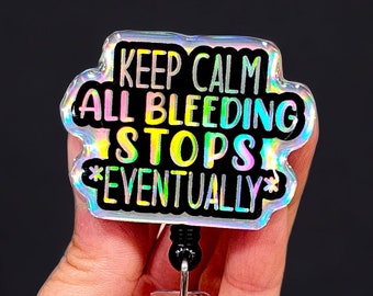 Può includere: Un porta badge olografico con la scritta "KEEP CALM ALL BLEEDING STOPS *EVENTUALLY*". Il badge ha un cordino retrattile in plastica trasparente e una clip in metallo. Il testo è in nero, bianco e vari colori.