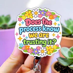 May include: A round badge reel with the text "Does the process know we are trusting it?" in colorful lettering. The badge is decorated with a floral border and attached to a clear plastic reel with a metal clip.