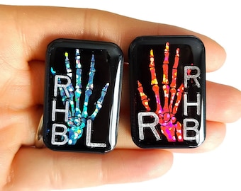 Può includere: Due piccoli portachiavi quadrati in resina nera con un design a mano scheletrica. Un portachiavi ha una mano scheletrica iridescente blu con le lettere RHB L stampate sul lato. L'altro portachiavi ha una mano scheletrica iridescente rossa con le lettere RHB R stampate sul lato.