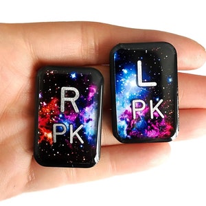 May include: Two black rectangular keychains with a galaxy print background. The left keychain has the letters "L PK" in white and the right keychain has the letters "R PK" in white.