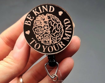 May include: A black and rose gold round badge with a brain and flower design and the text "Be Kind To Your Mind". The badge is attached to a black retractable badge reel with a clear plastic clip.