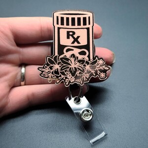 May include: A pink and black wooden badge reel with a floral design and a pill bottle with the letter "Rx" on it.
