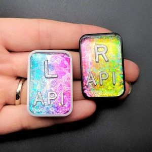 May include: Two resin keychains with a rainbow glitter background. One keychain says "L API" and the other says "R API".