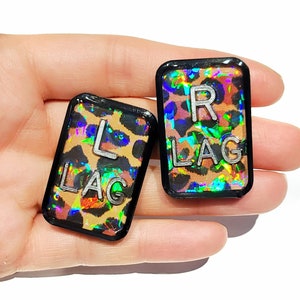 May include: Two black rectangular buttons with a leopard print background and iridescent finish. The buttons have silver lettering that reads "L LAG" and "R LAG".