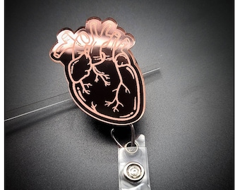 Puede incluir: Un portagafete en forma de corazón de color oro rosa con un diseño anatómico detallado del corazón. El portagafete tiene un carrete retráctil de plástico transparente.