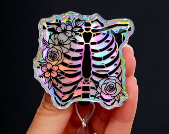 Pode incluir: Um carretel de crachá holográfico iridescente com uma imagem em preto e branco de uma caixa torácica com flores. O carretel está preso a um suporte de crachá retrátil de plástico transparente.