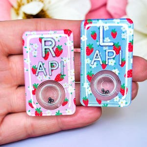 May include: Two rectangular, decorative pins. One is pink with strawberries, white flowers, and the letters "RAPI". The other is blue with strawberries, white flowers, and the letters "LAPI". Each pin has a clear, circular center with small black beads.