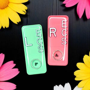 May include: Two rectangular, colorful cases, one green and one pink, each with the letters "LECS" and "RCS" in silver. Each case has a silver button and a small silver design. The cases are surrounded by pink, yellow, and white flowers.