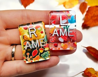 May include: Two small glass tiles with colourful autumn leaf designs and the words "RAME" and "LAME" printed on them.